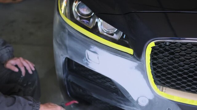 Auto body technician using compressed air gun to remove sanding dust and fine particles from primed front bumper before repainting luxury vehicle in workshop.