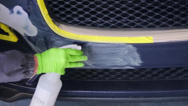 Auto technician spraying degreaser on repaired car bumper close-up in body shop, cleaning prepared surface before primer coating and automotive repainting. Degreasing of car surfaces, car painting.