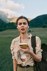 Fototapeta premium Woman traveler outdoors with backpack holds smartphone, surprised expression, mountain meadow backdrop, casual rugged style, candid lifestyle moment