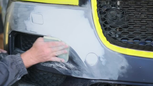 Auto body repair technician manually smoothing primed bumper surface with sanding pad before automotive refinishing and repaint coating process in garage. Car Surface smoothing before painting