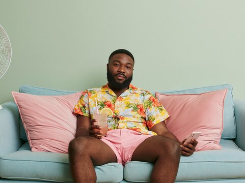 Black man wearing floral shirt and pink shorts sits casually on a pastel blue couch, holding a drink and smartphone, with a fan in the background