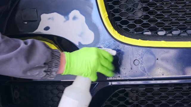Auto technician spraying cleaner on sanded car bumper close-up in repair workshop, removing dust and grease before primer coating and repainting process.