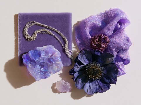 Overhead flat lay of purple fabric, a silver necklace, a large crystal, and a dark flower arranged on a pure white background for material mood board inspiration