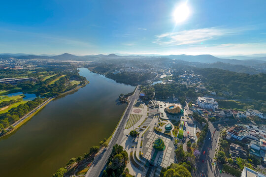 Aerial view: Dalat, Vietnam, featuring Xuan Huong Lake, Lam Vien Square, and city sprawl amidst rolling mountains on a bright day
