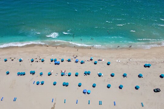 Aerial view of Singer Island beach featuring turquoise ocean waves, sandy shore, and rows of blue beach umbrellas with people in Riviera Beach, Florida, United States.