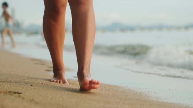 Bare feet walking along sandy shore, soft waves lapping ankles, solo traveler strolling past distant swimmers, warm sunlight casting long shadows, footprints imprinting then erased by tide, slow