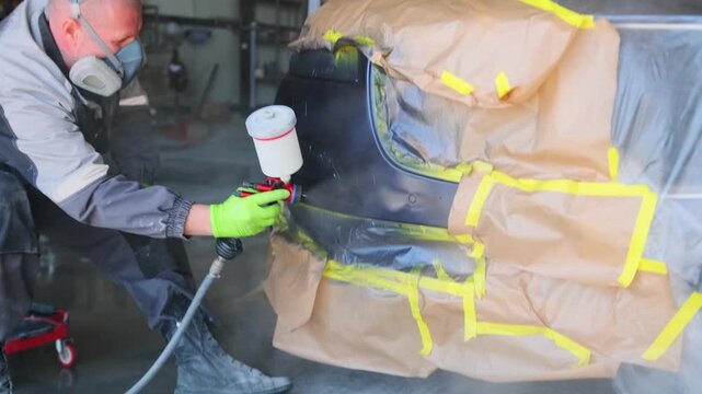 Professional auto body technician spraying protective clear coat lacquer on repaired vehicle bumper during refinishing process inside modern collision repair garage. car paint varnished painting car