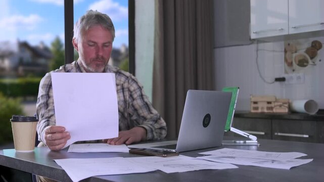 Engineer reviewing technical documents at kitchen table with laptop and construction paperwork, managing residential project calculations and remote planning tasks.