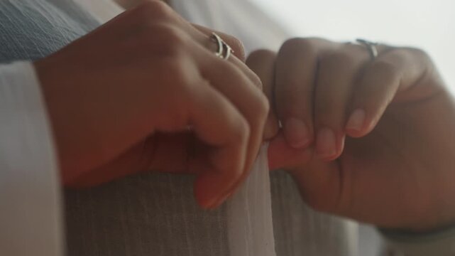 Hands fastening sheer blouse close up, confident morning prep with woman adjusting outfit before outing, subtle reveal of neckline under linen, warm backlight highlights movement of fingers securing