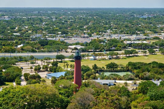 Aerial view of the red Jupiter Inlet Lighthouse surrounded by lush green trees and a park area with tennis courts in Jupiter, Florida, United States.