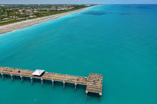 Aerial view of the Juno Beach Pier stretching over turquoise Atlantic waters toward a sandy coastline with beachgoers and coastal buildings Juno Beach, Florida, United States.
