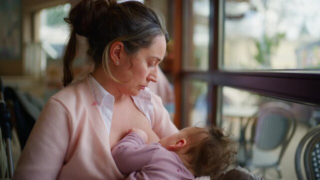 A mother in a light pink cardigan breastfeeds her baby while sitting beside a large window in a cafe