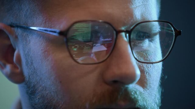 Close up shot of male Caucasian hacker wearing eyeglasses with reflection of programming codes working on computer indoors
