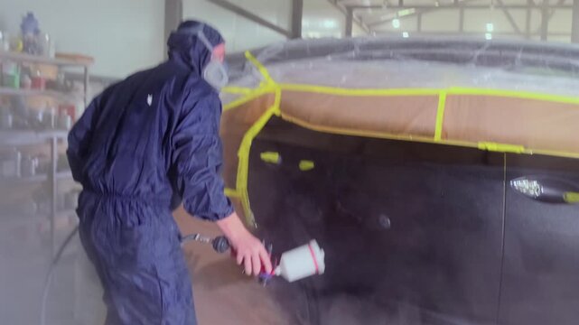 Professional auto body painter spraying glossy clear coat lacquer on repaired black vehicle door during automotive refinishing and collision repair process inside workshop.