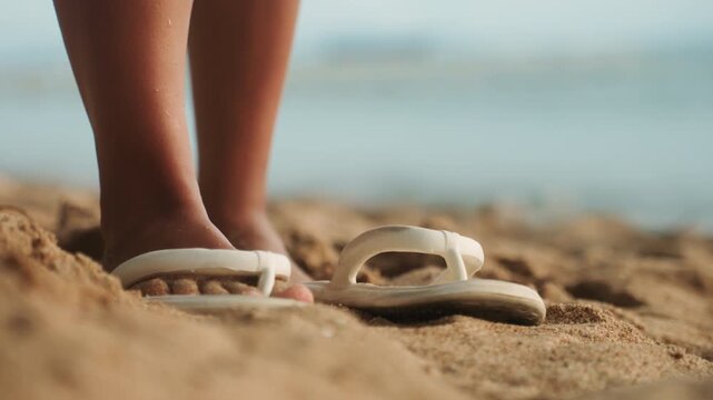 Flipflops on sandy beach closeup, young woman stands with toes sunk in warm grainy sand, white sandals cast soft shadow, blurred ocean horizon and gentle waves in background, golden light evokes