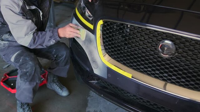 Auto body technician sanding repaired front bumper surface before primer and repaint application during vehicle restoration and automotive refinishing work in garage.