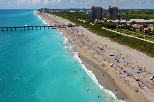 Aerial view of Juno Beach Pier stretching into the turquoise Atlantic Ocean with people on the sandy beach and coastal buildings in Juno Beach, Florida, United States.