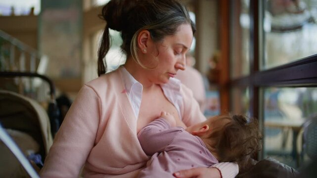 A mother in a light pink cardigan breastfeeds her baby while sitting beside a large window in a cafe