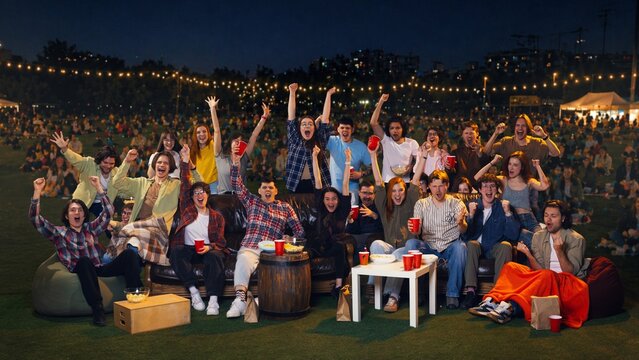 Young crowd raising hands at night fan zone watch party. Concept of sports viewing, victory reaction, public fan event, outdoor entertainment and group celebration.