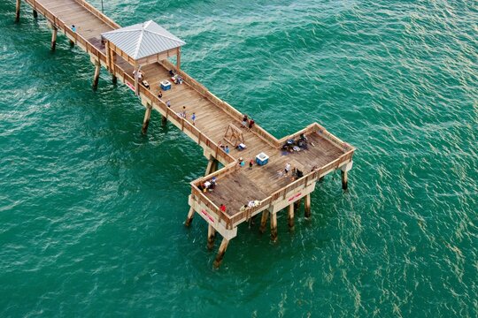 Aerial view of the T-shaped Deerfield Beach International Fishing Pier extending over turquoise ocean water with people fishing and walking in Deerfield Beach, Florida, United States.