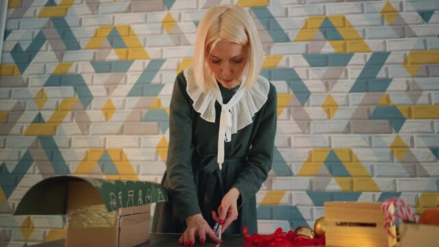 White woman wrapping gifts on desk, mosaic wall backdrop, cardboard box, red ribbon and scissors, warm lamp glow and string lights, focused artisan preparing holiday orders, tidy tabletop with bows
