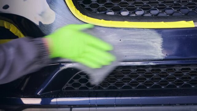 Auto technician spreading body filler on sanded car bumper close-up in repair garage, leveling damaged surface before primer coating and repainting work. Body filler application