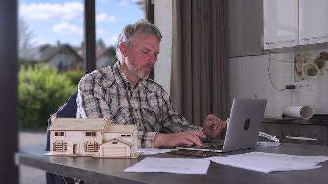 Engineer working on laptop at kitchen table with house model and technical paperwork, managing residential project documentation and remote construction planning.