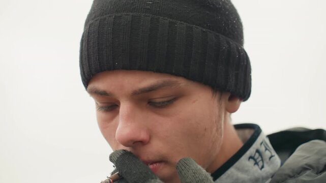 Closeup white man lighting cigarette, beanie and gloves framing face, fingers near lips as spark ignites, soft bokeh sky and distant city blur, facial details skin texture and light stubble visible,