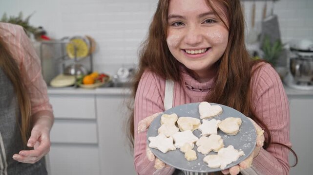 Smiling teenage girl showing heart shaped raw cookies on a plate in kitchen. Happy young baker with flour on face, family cooking together, domestic lifestyle, homemade pastry vlog
