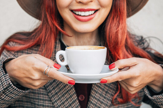 Woman with red hair and red nails holding a white cup of hot coffee, smiling and drinking a beverage