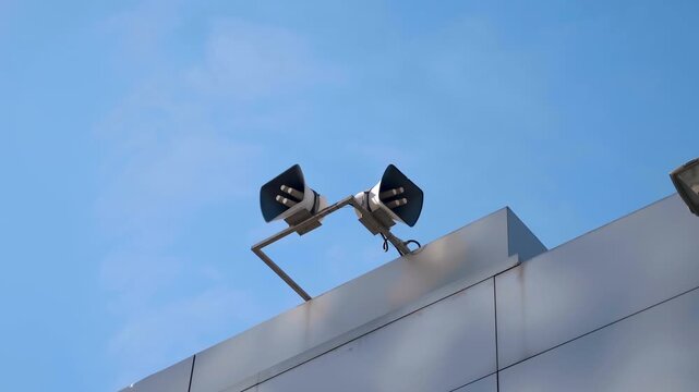 Two powerful loudspeakers are positioned high on the edge of a building against a blue sky. Shot in motion   