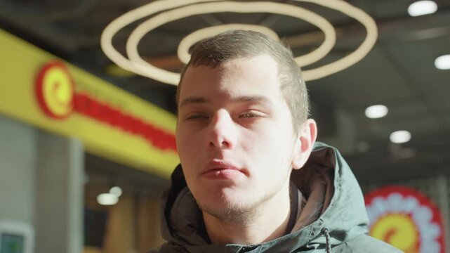 White man under halo lights, contemplative urban portrait with warm retail signage, indoor ring lighting, winter jacket hood, soft bokeh and solemn expression captured in closeup.