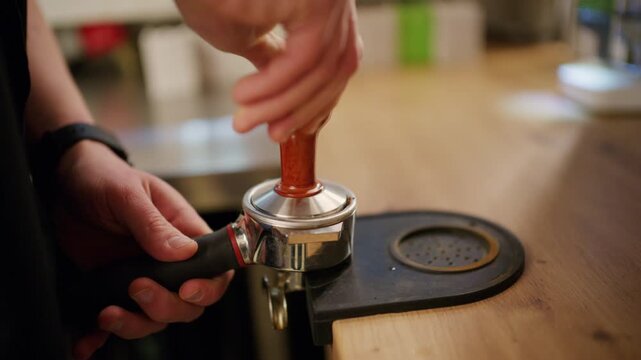 Close up of a barista pressing coffee grounds with a metal tamper inside a portafilter on a wooden cafe counter