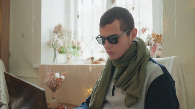 Young caucasian man coding on laptop at window, wearing green scarf and sunglasses, warm fairy lights and floral vase in background, focused remote developer debugging and typing in cozy study