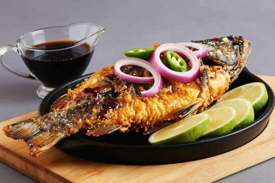 Whole fried bangus fish dish served with soy sauce. Crispy battered seafood garnished with red onion rings, chili slices, and lime wedges. Delicious meal presented in a black skillet.