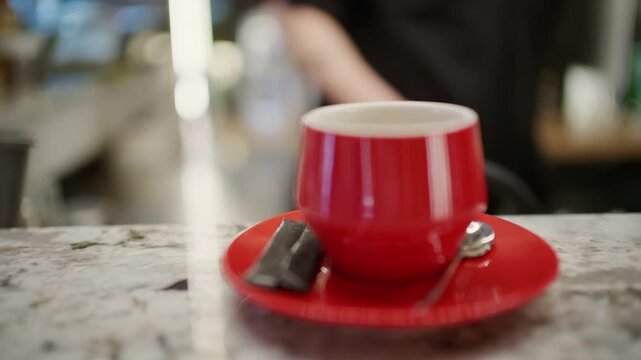 A red ceramic espresso cup on a matching red saucer is placed on a marble cafe counter with a small metal spoon beside it
