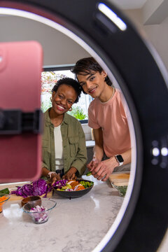 Couple preparing salad in kitchen while recording with ring light, pink phone, cutting board