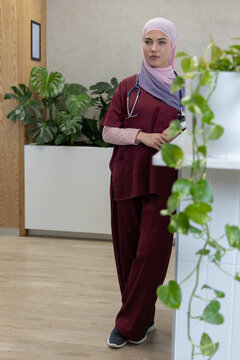 Female healthcare professional standing at clinic desk wearing maroon scrubs and hijab, stethoscope