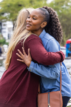 Diverse female friends hugging by pond in urban park wearing denim jacket and brown crossbody bag