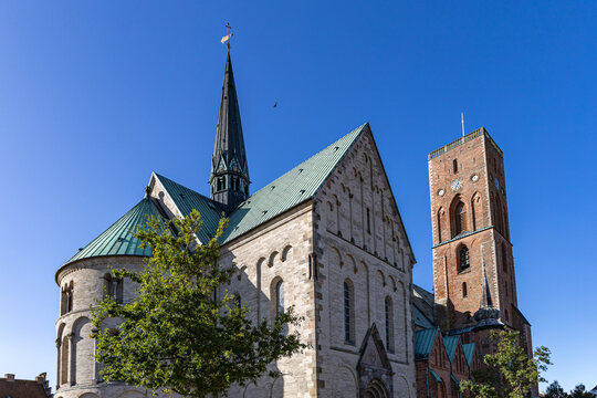 View of Ribe Cathedral featuring its Romanesque stone walls, green copper roofs, and the tall red brick Commoners' Tower under a clear blue sky in Ribe, Denmark.