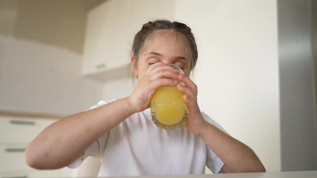 Cute girl drinking fresh orange juice from glass in bright kitchen. Thirsty child holding large glass. Smiling young girl enjoys healthy juice morning routine. Happy kid loves drinking beverage.