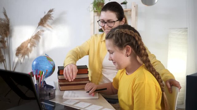 Mother helps daughter with homework. Happy mother and young daughter look at laptop screen. Child writes in notebook during home study. Family values school education. Laptop displays homework.