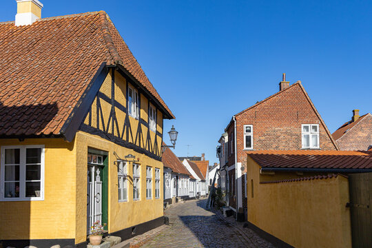 View of historic half-timbered houses with yellow facades and red tiled roofs along a narrow cobblestone street under a clear blue sky in Ribe, Denmark.