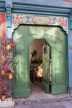 View of an ornate historic green wooden door with carved angels and inscriptions leading to a courtyard with a wooden cart Ribe, Denmark.