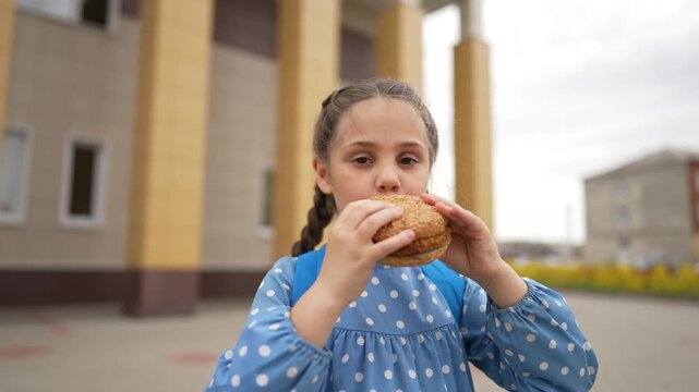 Schoolgirl with braids eats sesame bun outside school. Hungry pupil bites tasty bread during lunch break. Student with backpack enjoys snack near school building. Child chews bun outdoors.