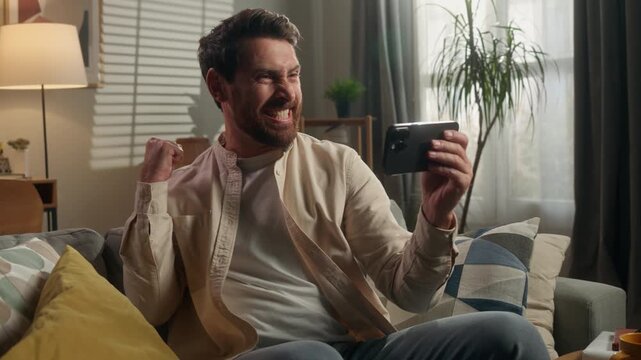 Happy Caucasian middle-aged man looking in mobile phone winning yelling joyful winner adult male watching sport football match with smartphone celebrate win betting achieve bet success yell at home