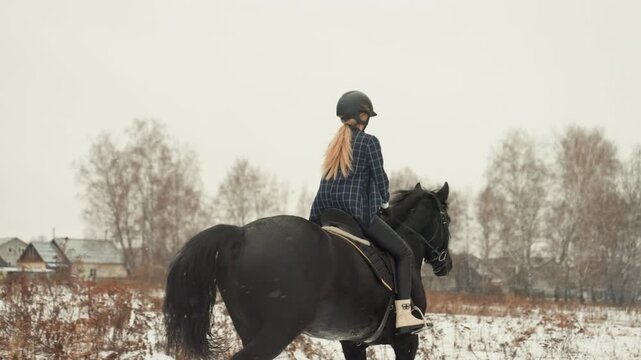 Caucasian rider on black horse strolling through snowdusted meadow near village, helmeted profile with braid practicing quiet posture and gentle canter during equestrian education moment falling snow