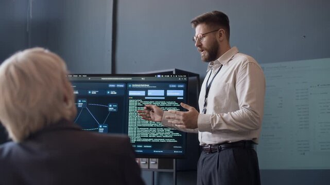 Low angle view of confident male Caucasian network engineer giving presentation with programming codes on screen and talking to female colleague in office