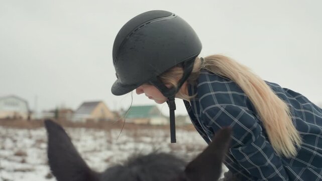White rider smiles over horse ears in snowy field, joyful tandem bonding and preride connection, helmeted student checking reins with gentle touch, village houses in background, warm expression