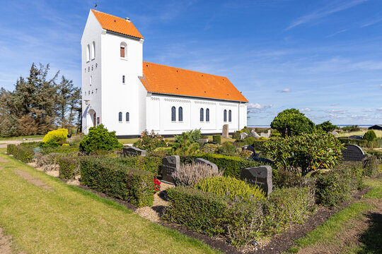 View of Haurvig Kirke with its white facade and orange-tiled roof surrounded by a well-kept churchyard under a clear blue sky in Hvide Sande, Denmark.
